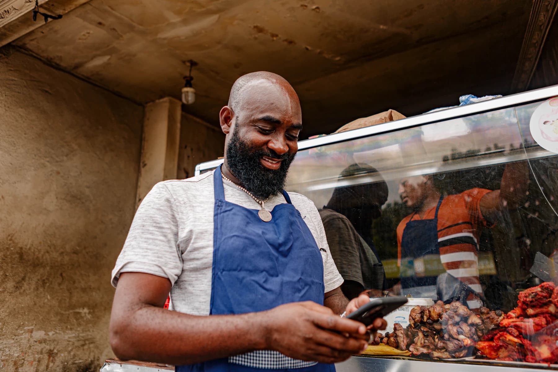 A street-food vendor in a blue apron reads a message on his phone beside his glass counter